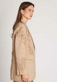 Sacou bej cu broderie florală complicată, accente texturate și detalii cu nasturi pe mâneci, prezentat dintr-o vedere laterală.