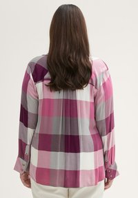 Kvinde med langt brunt hår iført en pink, lilla og gråternet bluse med lange ærmer og lyse bukser, vist fra ryggen.