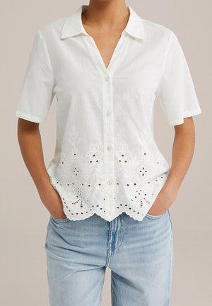Kvinde iført en hvid kortærmet knapbluse med blomsterudskæringsbroderi, kombineret med lyseblå jeans, hænderne i lommerne.