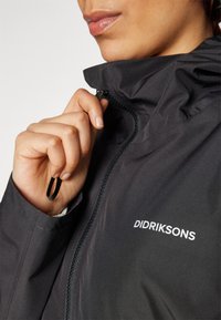 Giacca nera impermeabile con chiusura a zip, colletto alto, coulisse regolabile e logo bianco sulla parte anteriore. Textura morbida.