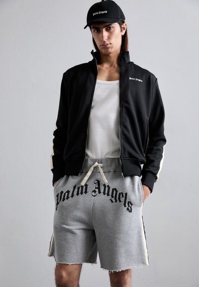 Male wearing black zip-up jacket and cap, white tank top, and gray shorts with "Palm Angels" print, standing against a plain light background.