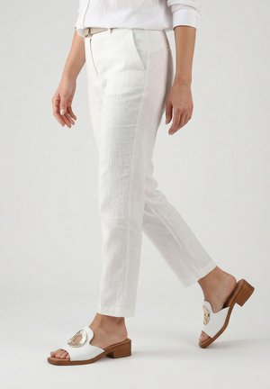 Kvinde iført hvide croppede bukser og hvide hælede sandaler med gyldne dekorative accenter, der går mod en ensfarvet baggrund.