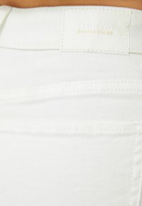 Primer plano de tela de mezclilla blanca con costuras visibles y una etiqueta rectangular cosida que dice "Denim Bershka Est.98" en la cintura.