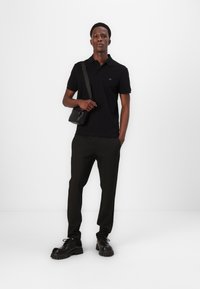 Camisa polo preta com um pequeno logotipo, combinada com calças pretas risca de giz e sapatos pretos robustos. Modelo segurando uma pequena bolsa preta.