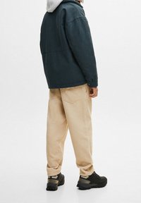 Person som bär beiga manchesterbyxor, svarta vandringsskor och en mörk jacka med grå huva, stående mot en enfärgad vit bakgrund.