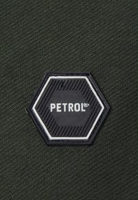Parchment de goma negro con forma hexagonal que presenta el texto "PETROL" en relieve sobre un fondo de tejido verde oscuro texturizado.