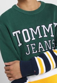 Pull vert en tricot avec l'inscription "TOMMY JEANS" en lettres blanches, manches rayées en bleu marine, jaune et blanc. Finition en tissu texturé.
