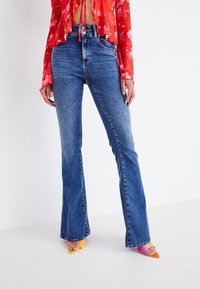 Mujer vestida con unos vaqueros de campana azules, una blusa corta de flores rojas y unos zapatos de tacón de punta coloridos, de pie contra un fondo blanco.