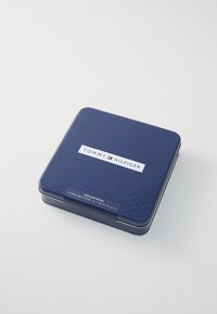 Boîte en métal carrée de couleur bleu marine avec un motif en damier texturé, arborant une étiquette blanche avec la marque "TOMMY HILFIGER" et les détails du produit.