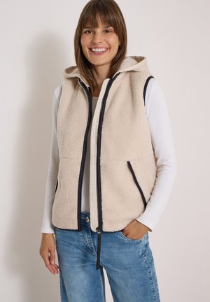 Femme souriante portant un gilet zippé en polaire beige avec une bordure noire, une chemise blanche à manches longues et un jean bleu, debout devant un fond uni.