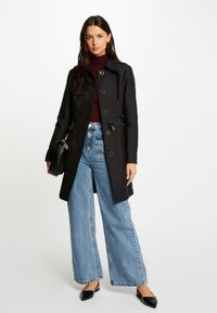 Zwarte trenchcoat met een ceintuur om de taille, knoopsluiting en zijzakken. Gecombineerd met lichtblauwe jeans met wijde pijpen en zwarte puntige flats.