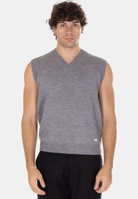 Giovane uomo che indossa un gilet grigio lavorato a maglia senza maniche e pantaloni neri, in piedi con un'espressione neutra contro uno sfondo semplice.