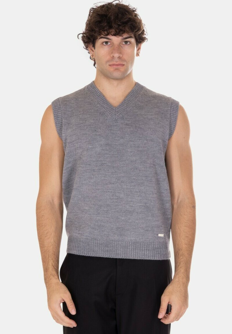 Giovane uomo che indossa un gilet grigio lavorato a maglia senza maniche e pantaloni neri, in piedi con un'espressione neutra contro uno sfondo semplice.