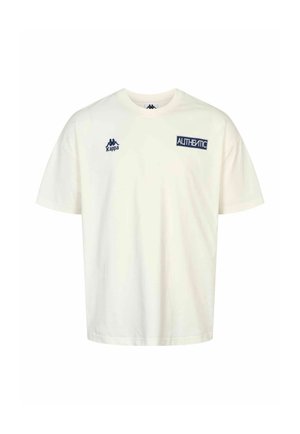Ung mand med kort krøllet hår og tatoverede arme bærer en hvid "Authentic" Kappa T-shirt og blå jeans, kigger til siden med hånd på nakken.
