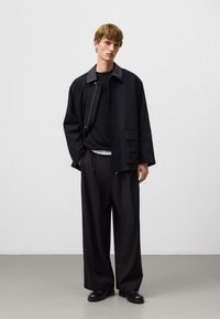 Czarna kurtka z kontrastowym kołnierzem, noszona na czarnym swetrze. Luźne czarne spodnie i czarne buty o minimalistycznym wzornictwie.