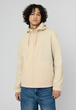 Veste légère beige avec capuche, fermeture éclair à l'avant et poignets élastiques. Le tissu a une texture lisse, avec un design minimaliste.