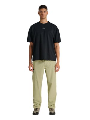 Ung mand står, iført en sort T-shirt med HALO-logo, beige cargobukser med lynlåslomme og beige vandresko.