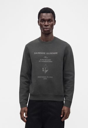 Un homme portant un sweat-shirt gris foncé avec du texte et un motif floral, debout sur un fond blanc uni.