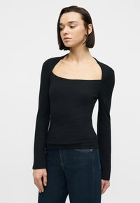 Νεαρή γυναίκα με ίσια, σκοτεινή καρέ κόμμωση φοράει μαύρο τοπ με μακριά μανίκια και τετράγωνο άνοιγμα στο ντεκολτέ, καθώς και σκούρο μπλε τζιν, μπροστά σε λευκό φόντο.