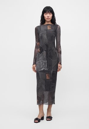 Femme aux longs cheveux noirs portant une robe midi ajustée à manches longues avec un imprimé patchwork denim noir et des talons ouverts noirs.