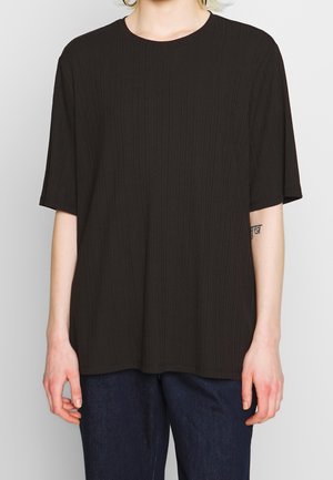 Person trägt ein schwarzes, kurzärmeliges, strukturiertes, locker sitzendes Hemd und dunkelblaue Hosen, steht vor einem schlichten hellen Hintergrund.