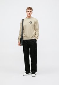 Beige kapucnis pulóver logóval, fekete cargo nadrág zsebekkel és fekete sportcipő. A modellen fekete válltáska van, sima háttér előtt áll.