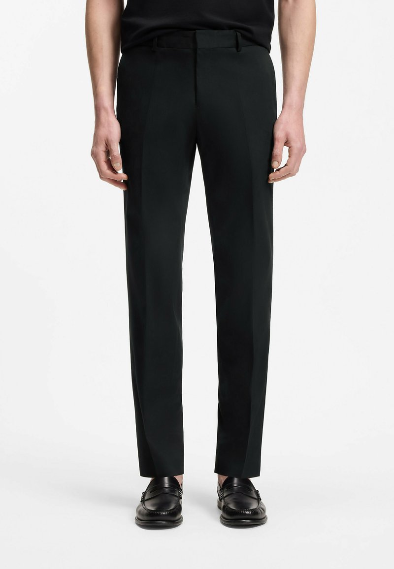 Homme portant un pantalon habillé noir slim-fit et des mocassins en cuir noirs, debout sur un fond blanc.