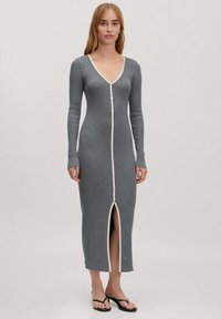 Langes graues ribbelndes Kleid mit tiefem V-Ausschnitt, durchgehender Knopfleiste und weißem Besatz. Mit langen Ärmeln und einem Schlitz vorne. Getragen mit schwarzen Flip-Flops.