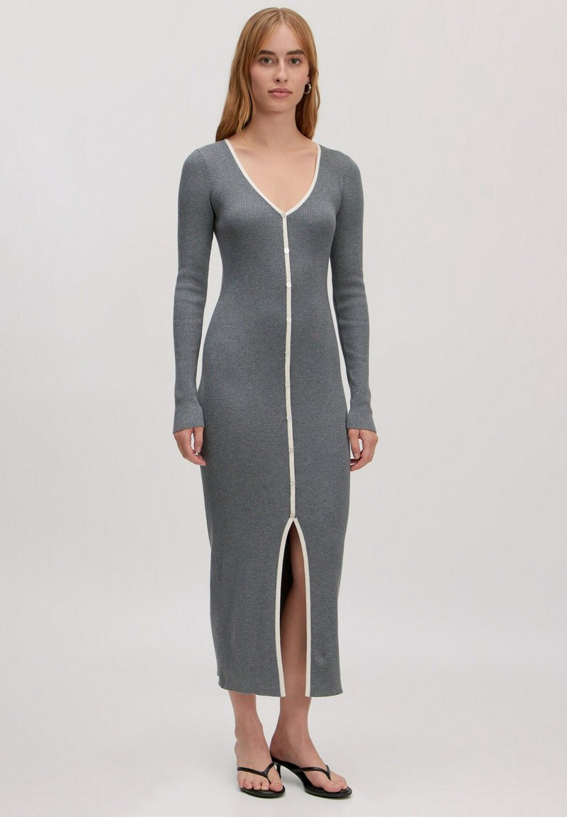 Langes graues ribbelndes Kleid mit tiefem V-Ausschnitt, durchgehender Knopfleiste und weißem Besatz. Mit langen Ärmeln und einem Schlitz vorne. Getragen mit schwarzen Flip-Flops.