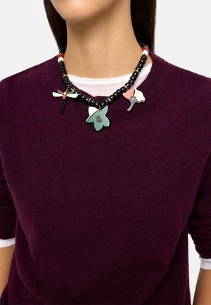 Naszyjnik z czarnych koralików z trzema dużymi, kolorowymi emaliowanymi zawieszkami w kształcie kwiatu i ważki, noszony na bordowym swetrze i białej koszuli.