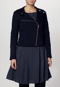 Veste courte noire avec une fermeture éclair diagonale, des manches texturées et des garnitures en cuivre, portée sur une robe marine à motifs avec des pois blancs.