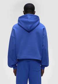 Felpa blu con cappuccio regolabile e vestibilità oversize. Presenta un tessuto testurizzato e maniche ampie, abbinata a pantaloni della tuta coordinati.