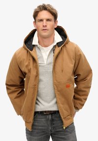 Chaqueta marrón con cremallera y capucha, con forro negro, grandes bolsillos frontales y una pequeña etiqueta naranja en el lado izquierdo.