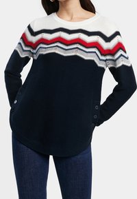 Navy gestrickter Pullover mit einem weißen, roten und blauen Zickzackmuster, rundem Ausschnitt und seitlicher Knopfdetails. Weiche Textur und lockere Passform.