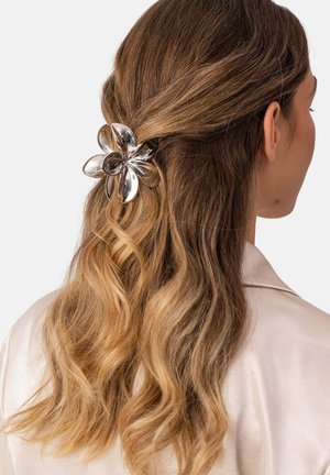Woman with long wavy blonde hair half pinned back by a shiny silver flower hair clip, wearing a light-colored shirt.
