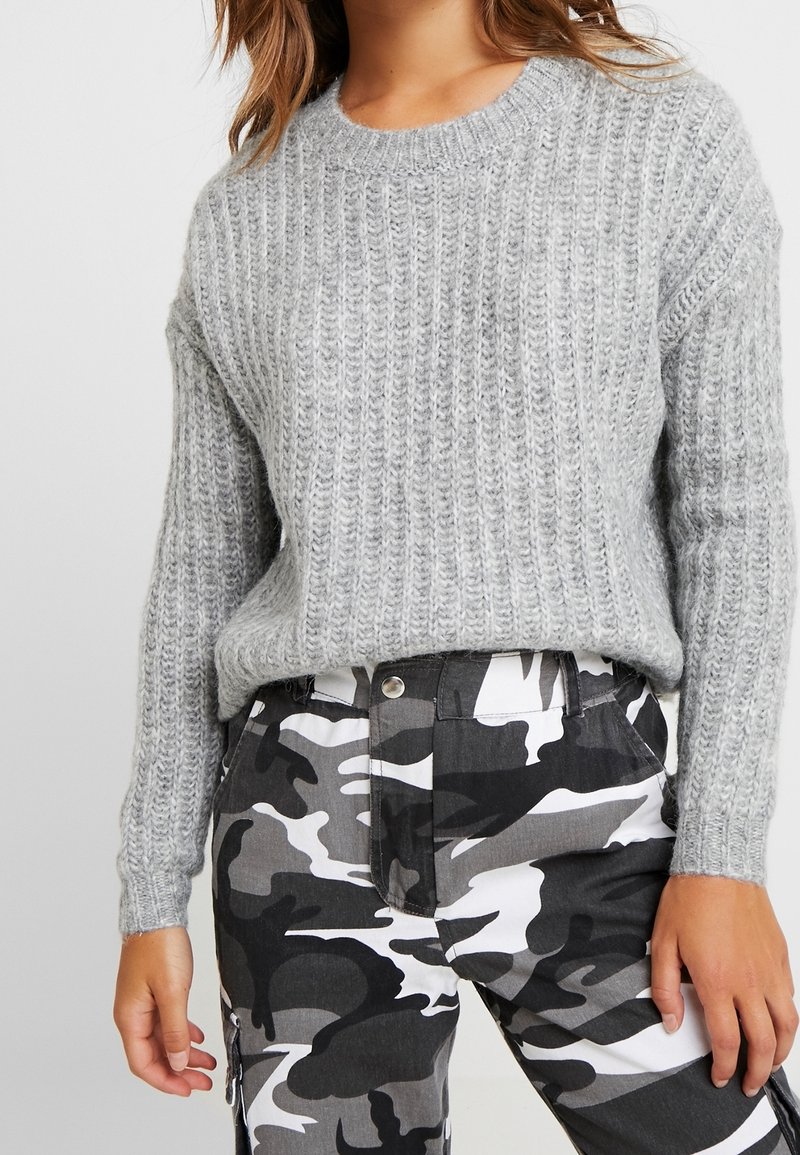 Femme portant un pull en tricot gris clair texturé et un pantalon camouflage noir, blanc et gris, debout les bras détendus le long du corps.