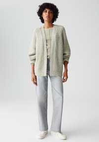 Lichtgroene, gestructureerde cardigan met verlaagde schouders, gedragen over een lichtbeige top, gecombineerd met lichtgrijze jeans en witte sneakers.