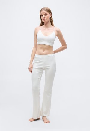 Mladá žena stojící bosá, oblečená v bílém krátkém tílku a bílých volných kalhotách na světlém jednobarevném pozadí.