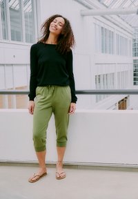 Kvinne med krøllete hår iført svart langermet topp, olivengrønne kuttede bukser og flip-flops som står innendørs med lukket øyne og smiler.