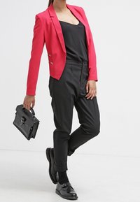 Blazer texturizado rojo con herrajes plateados, top negro, pantalones negros a medida y zapatos negros, sosteniendo un pequeño bolso negro.