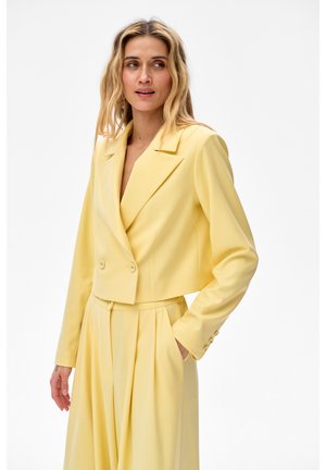 Kobieta w jasnożółtej, dopasowanej marynarce i pasujących, plisowanych spodniach z wysokim stanem, stojąca z jedną ręką w kieszeni.