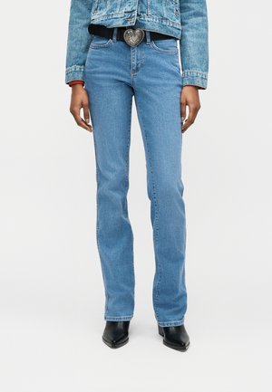 Donna che indossa jeans dritti a vita alta blu, cintura con fibbia a forma di cuore nera, giacca di jeans e stivali neri con punta a punta.