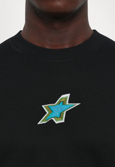 Dark-skinned person wearing a black crewneck shirt with a small embroidered geometric star design in blue, green, and white on the chest.