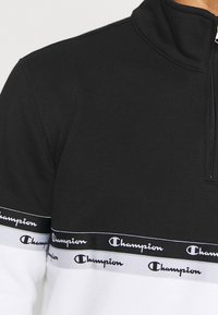Pullover a mezza zip in bianco e nero, realizzato in tessuto morbido. Presenta un logo Champion su una fascia orizzontale sul petto.