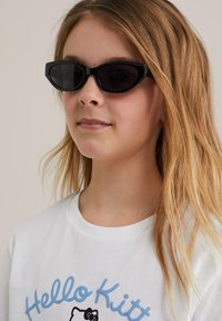 Gafas de sol estilo gato negras con lentes oscuros, usadas por una persona con cabello largo y castaño claro, combinadas con una camiseta blanca con un diseño gráfico azul.