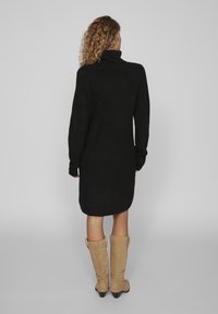 Abito nero lavorato a maglia con un colletto alto, maniche lunghe e una vestibilità over. Arriva a metà coscia. Indossato con stivali beige in camoscio al ginocchio.