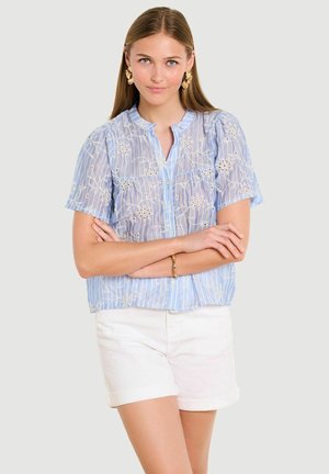 Mladá žena v modrej blúzke s kvetinovou výšivkou s krátkymi rukávmi a bielych kraťasoch stojí so založenými rukami.