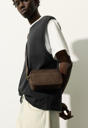 Persoană tânără purtând un top negru fără mâneci, cămașă albă, pantaloni deschiși și o geantă maro texturată purtată în diagonală.