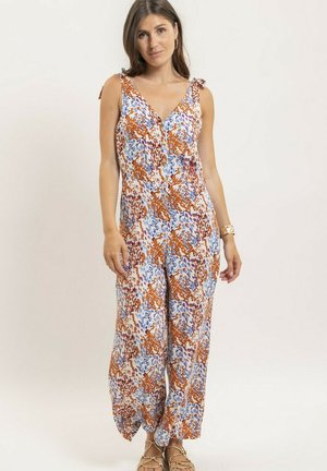 Femme portant une combinaison sans manches à jambes larges avec un motif floral abstrait orange, bleu et marron, debout devant un fond clair uni.