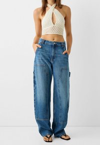 Bílý zkrácený halter top s texturovaným vzorem a výřezem ve tvaru klíče; modré široké džíny s volným střihem a bočními kapsami.